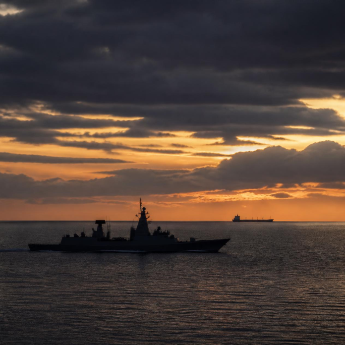 A naval warship and a commercial cargo vessel navigating open ocean waters, representing maritime security and seafarer protection.